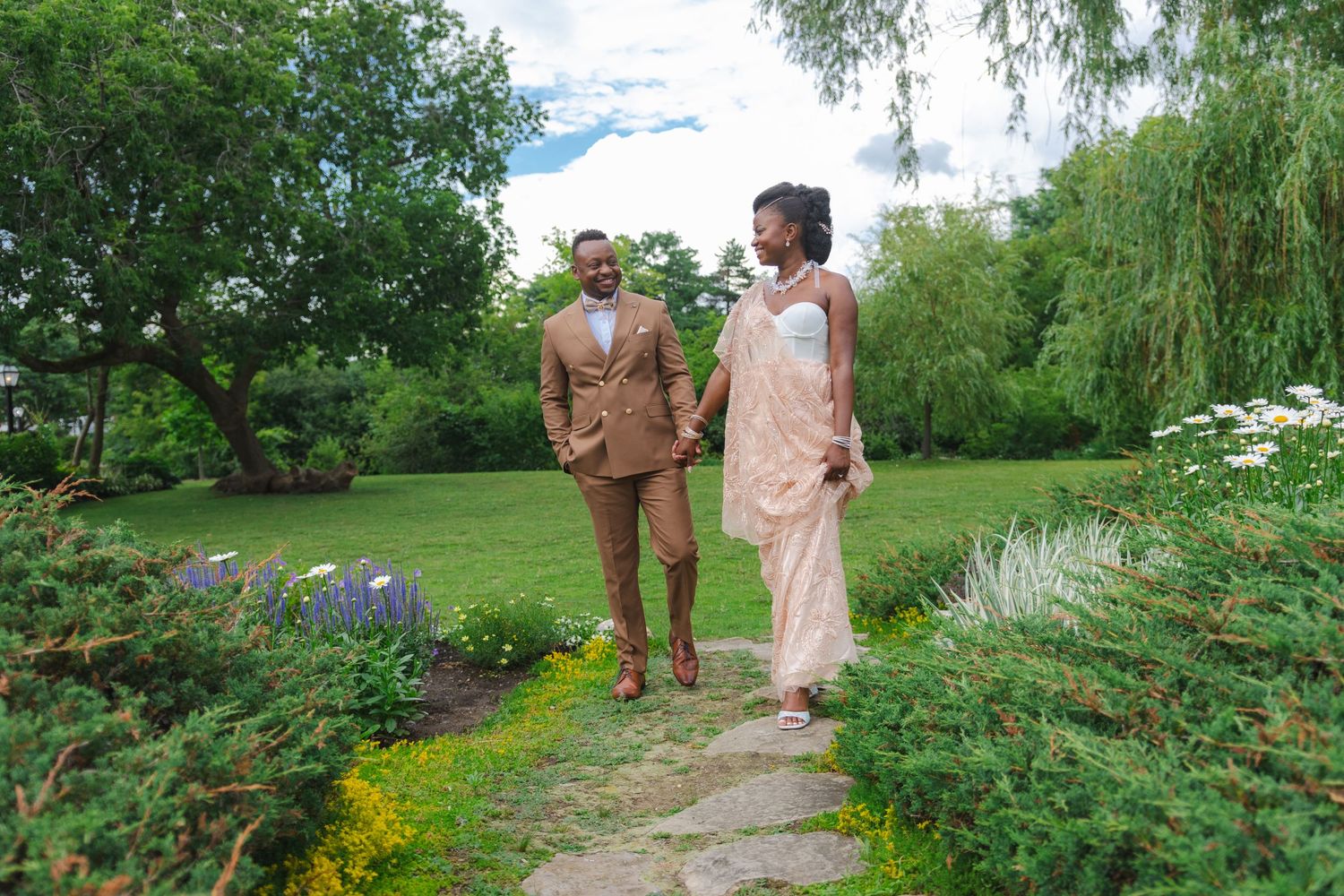 Two people walk together along a garden path surrounded by lush greenery and flowering plants on a sunny day.