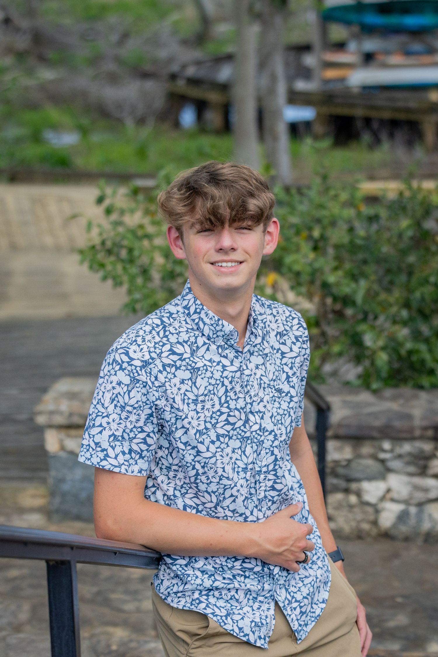 Young person in blue floral print shirt poses outdoors near stone wall and greenery.