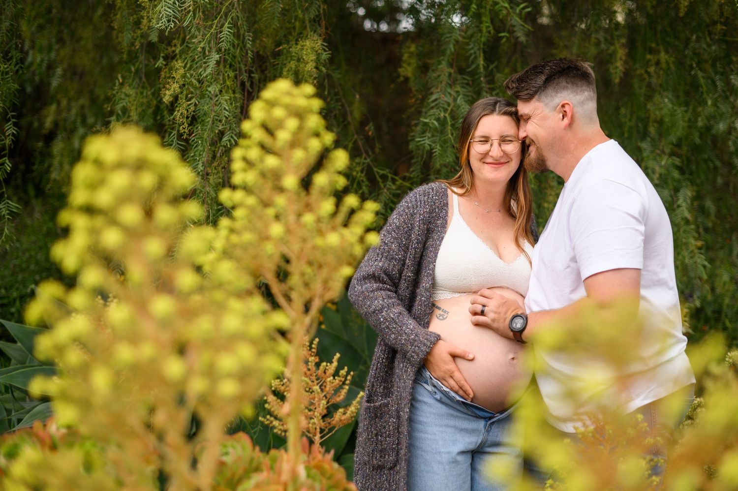 A couple shares an intimate moment in a garden setting with yellow wildflowers and weeping willow trees in the background.