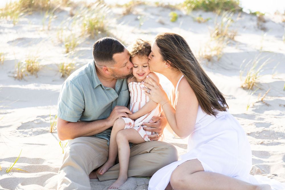 A happy family sitting on sandy beach, parents kissing their toddler on the cheeks in warm sunlight.