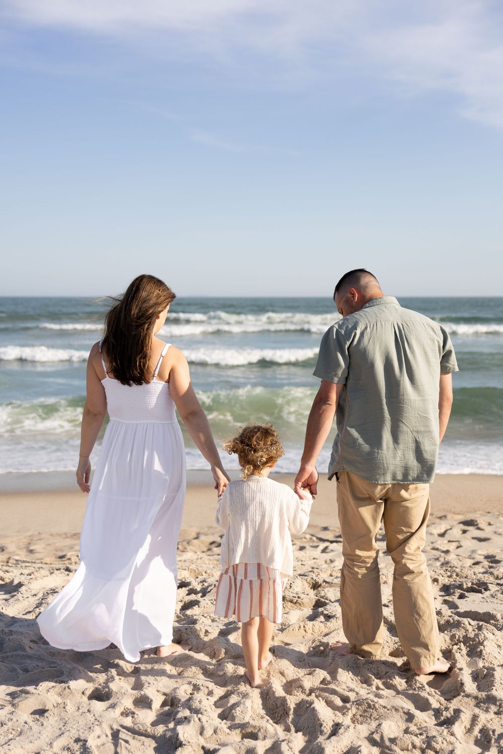 Family of three holding hands on sandy beach, parents and child facing ocean waves under blue sky.