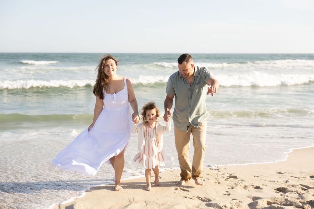 Family of three walking hand-in-hand on a sunny beach, parents swinging their toddler between them near ocean waves.