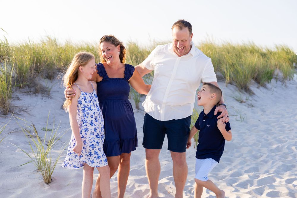 A happy family of four laughs together while walking on a sandy beach near grass dunes.