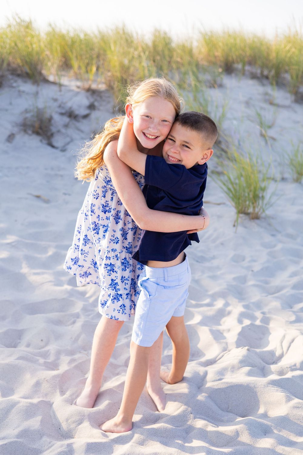 A smiling girl in a floral dress hugs her younger brother on a sandy beach with dunes.