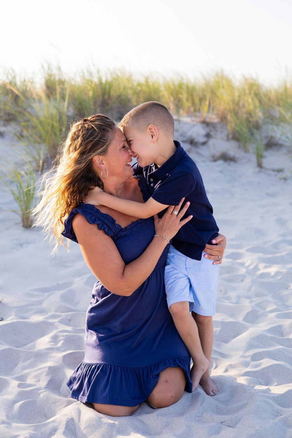 A mother kneeling on sandy beach dunes, hugging and smiling face-to-face with her young son.