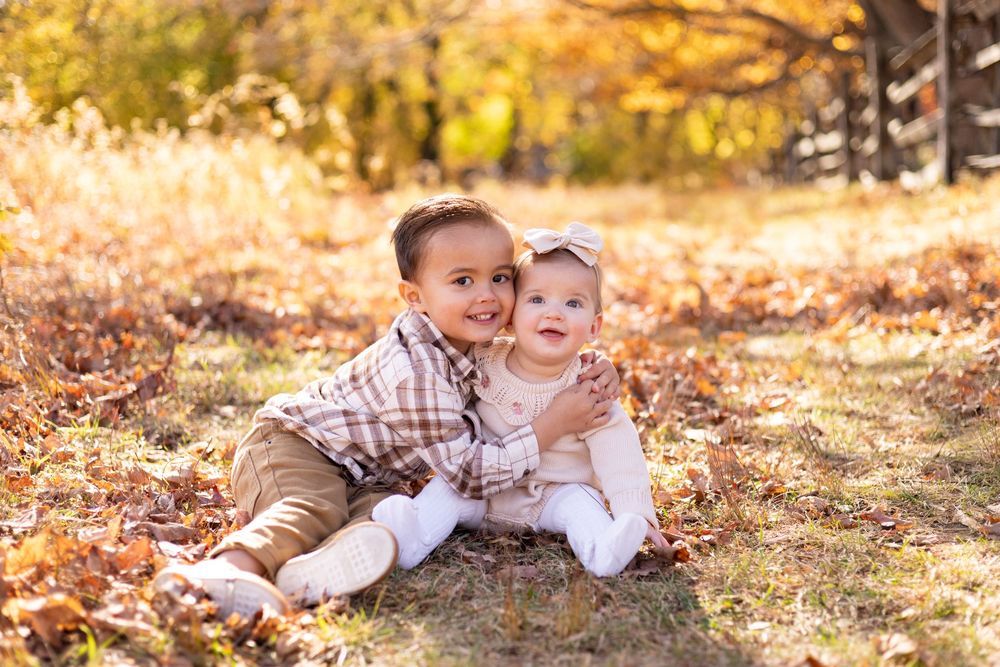 Two young siblings sit together in autumn leaves, smiling outdoors.