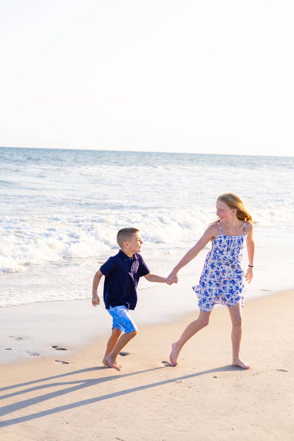 Two children holding hands and running along a sunny beach with ocean waves in the background.