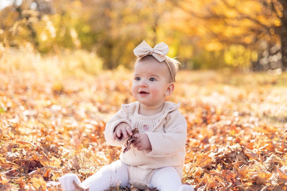 Adorable baby girl with bow headband sitting in fall leaves outdoors.