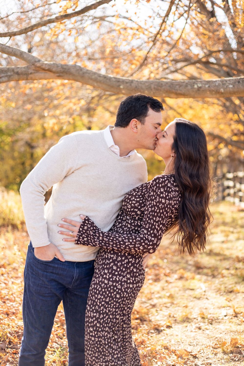 Couple sharing a romantic kiss outdoors beneath bare branches in autumn foliage.