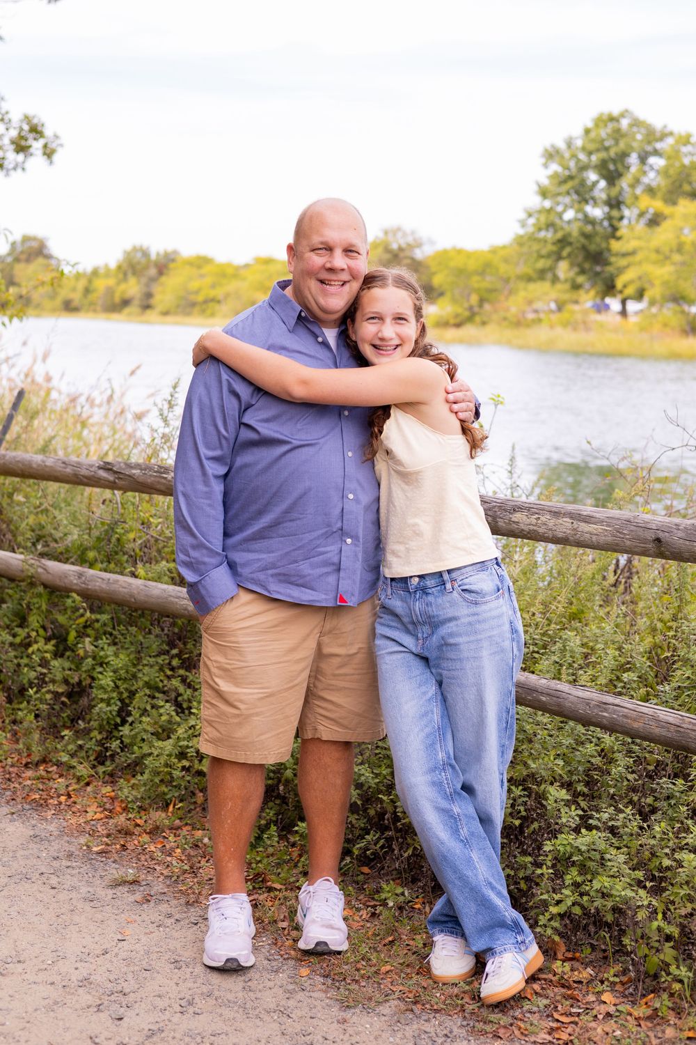 A bald man and young girl share a warm hug near a wooden fence by a scenic lake.