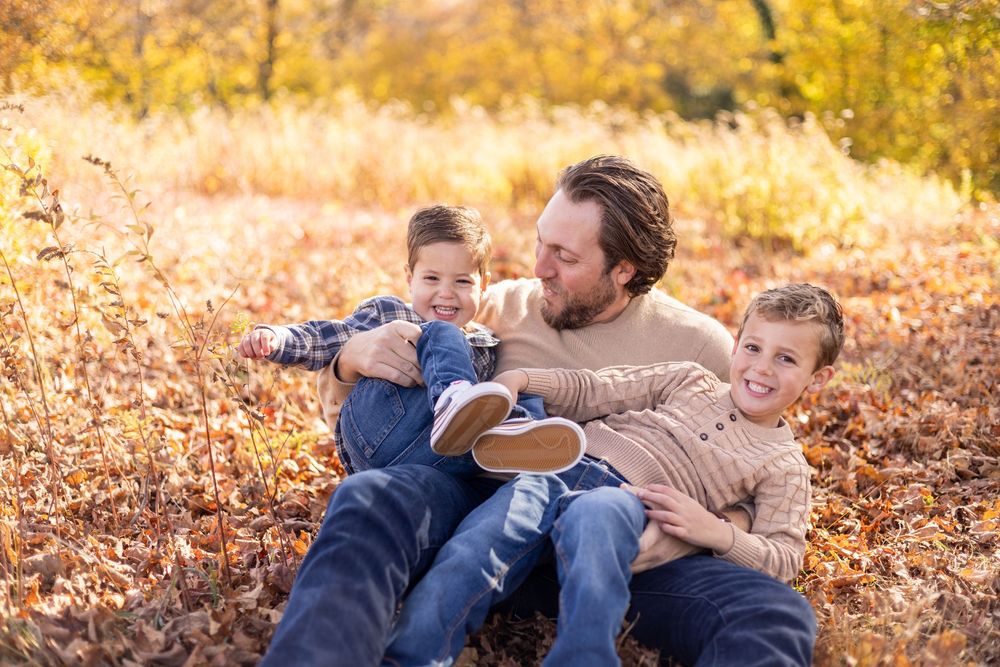 Father sitting outdoors in fall foliage laughing with two young children in his lap.