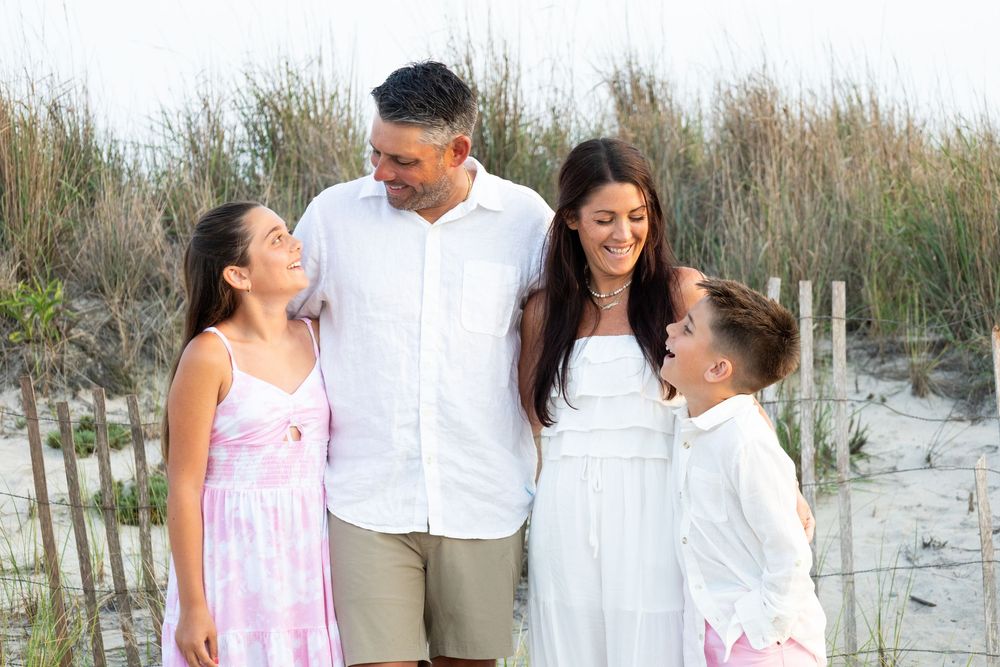 Smiling family of four poses together near beach dunes at sunset.