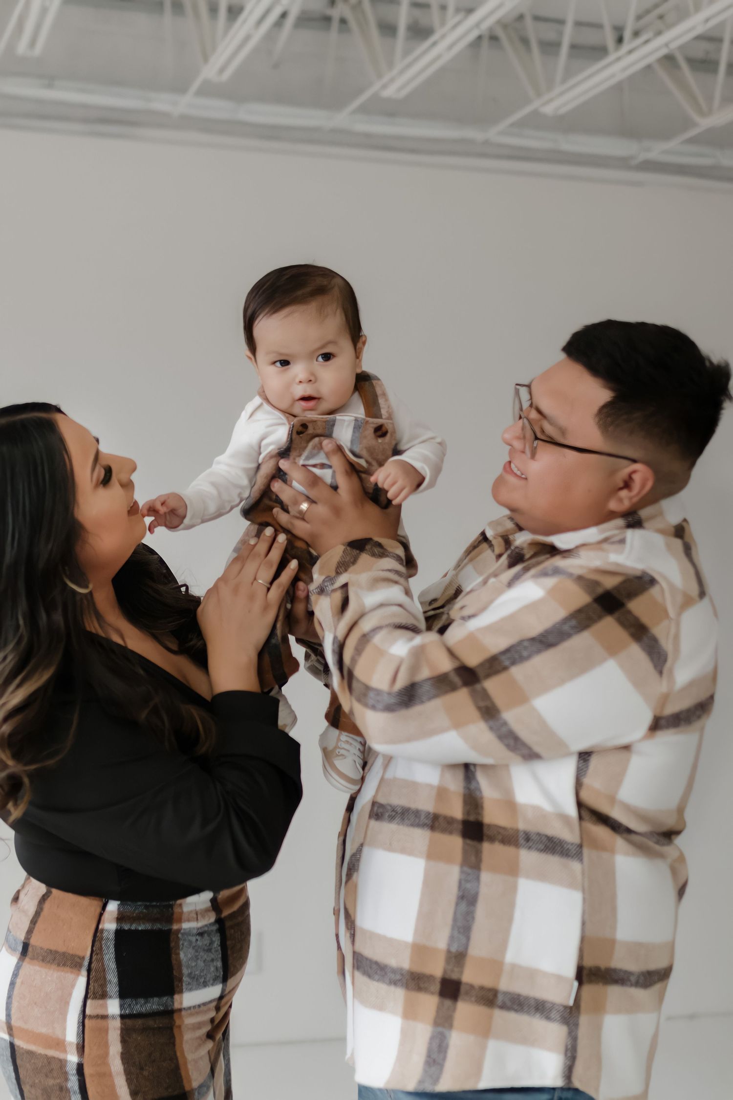 A loving family moment captured indoors against a white wall as parents lift their baby wearing matching plaid outfits.