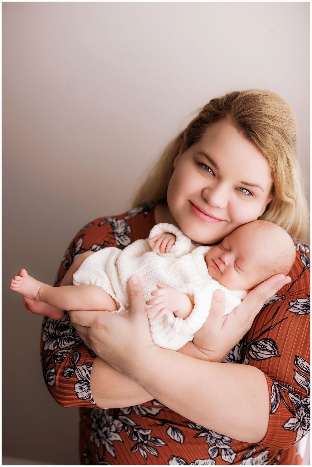 A tender portrait showing a sleeping newborn baby in white clothing being held against floral patterned fabric.