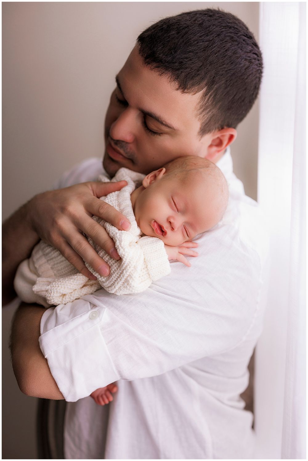 A tender moment shared between a baby sleeping peacefully in white against someone in a white shirt by a window.