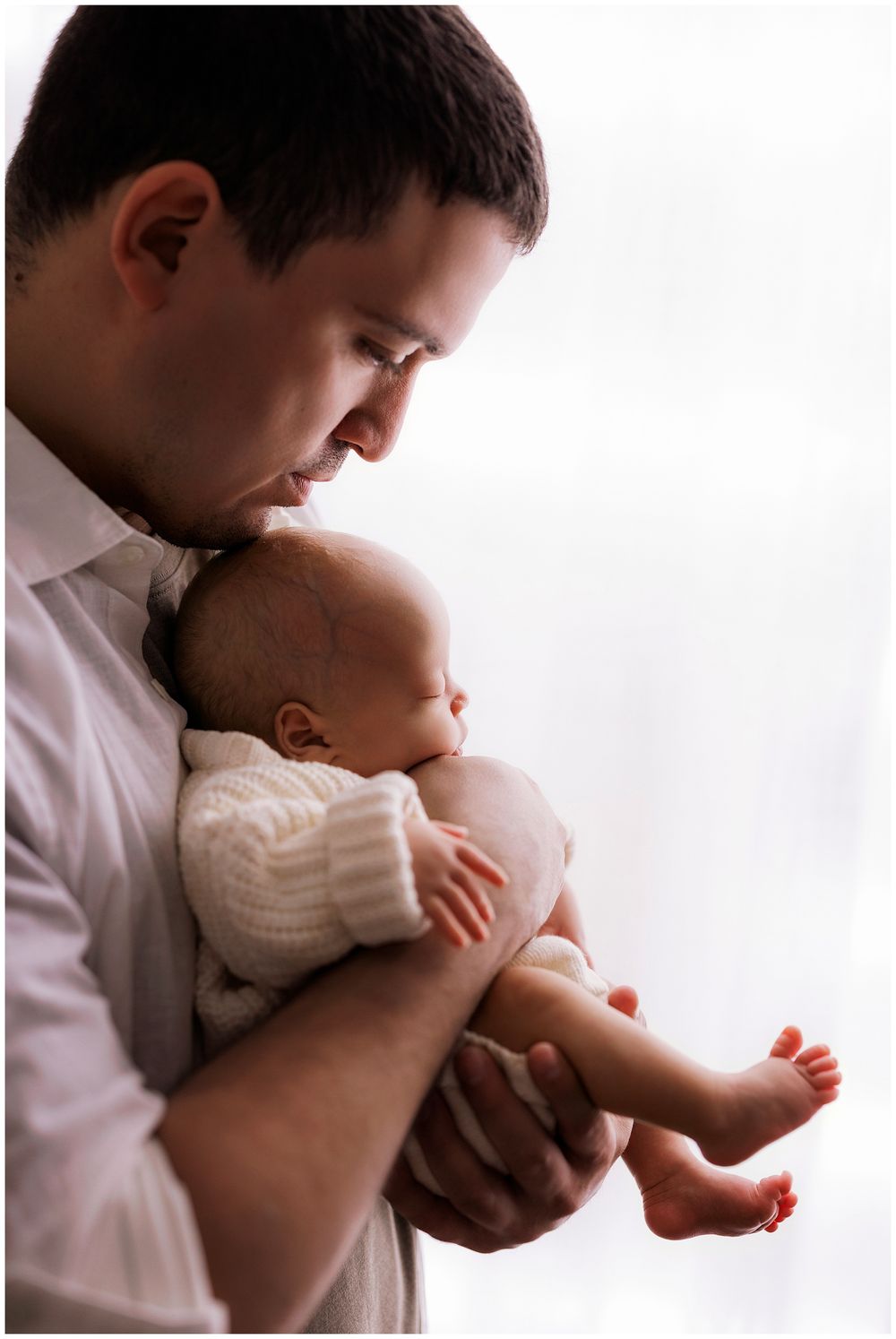 An adult in a white shirt holds and gazes lovingly at a sleeping baby wearing a white knit sweater by a bright window.