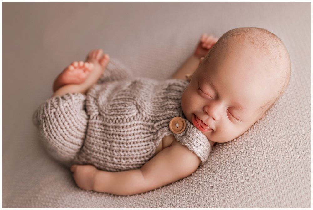 Sleeping newborn in cozy knit sweater lying on soft gray backdrop during professional photoshoot.
