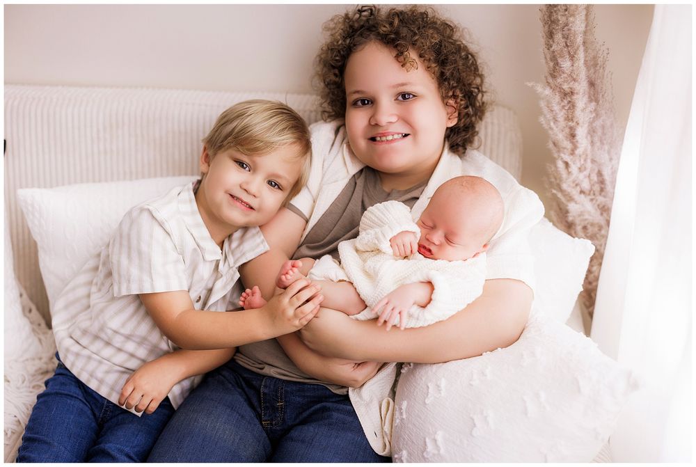 Three siblings share a tender moment together on a white couch with pampas grass decor in the background.