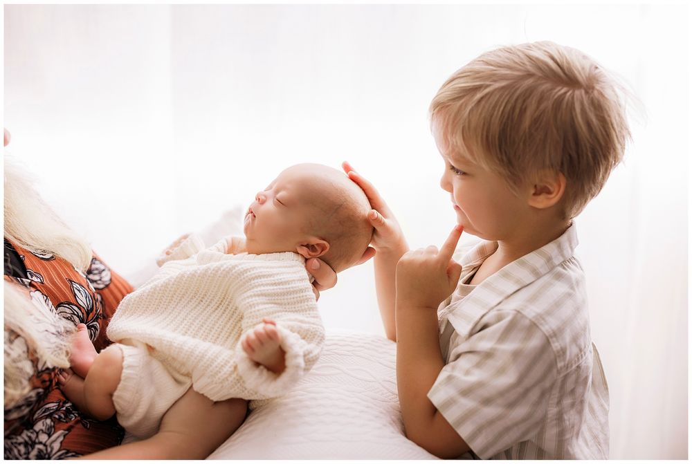 Toddler in striped shirt gently touching and looking at infant wearing white dress on white bedding.