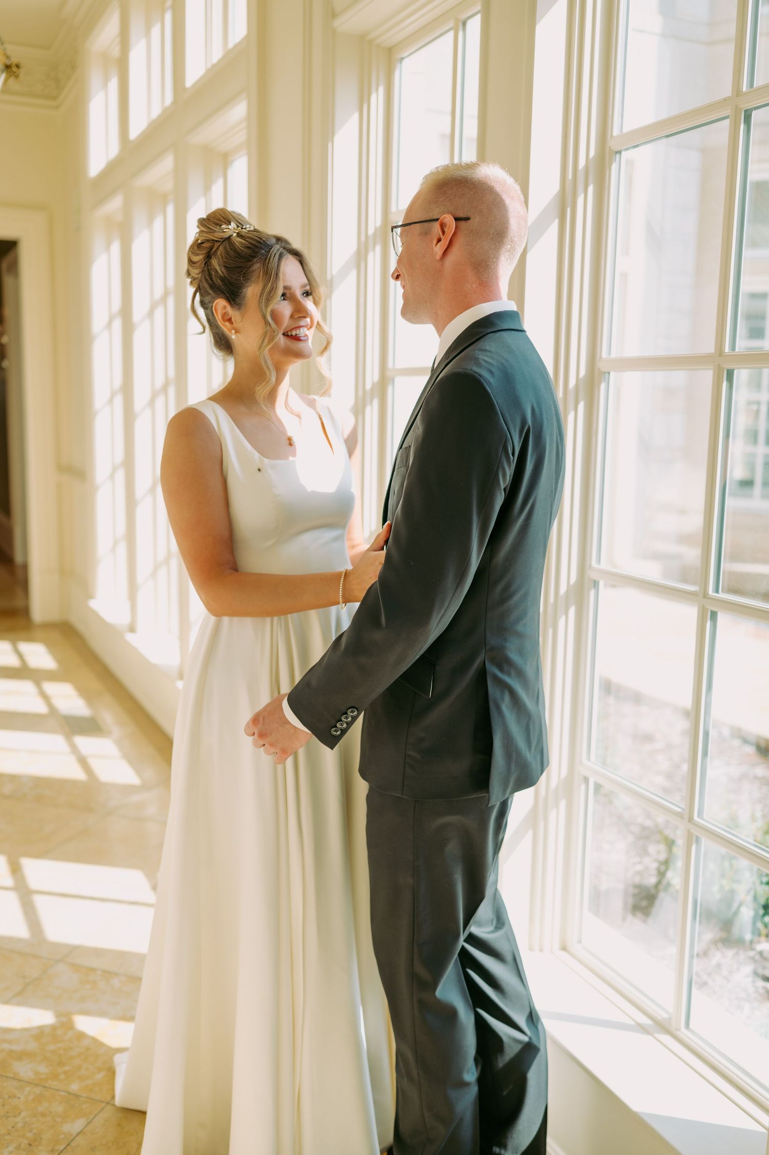 Sun-drenched wedding portrait of a couple sharing a joyful moment by large windows.