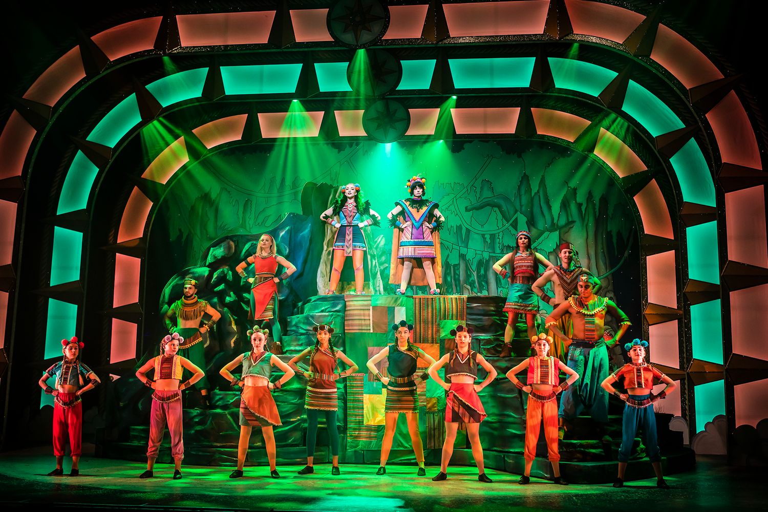 Musical theater stage with green archway lighting featuring performers in colorful costumes arranged in formation.