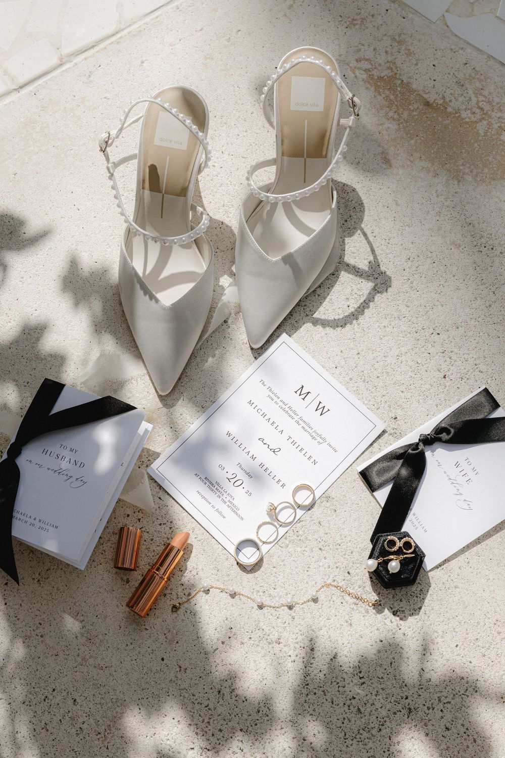 Elegant white bridal heels and wedding rings styled with invitation cards and accessories on a light concrete surface.
