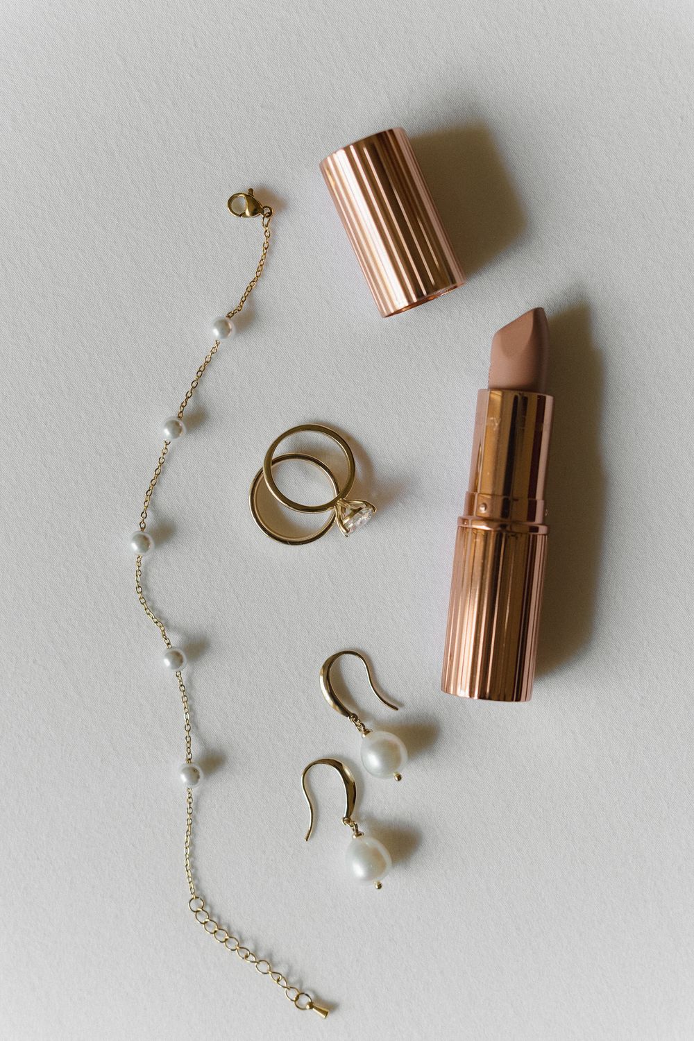 Feminine accessories including a rose gold lipstick, delicate gold jewelry, and pearl earrings arranged on white background.