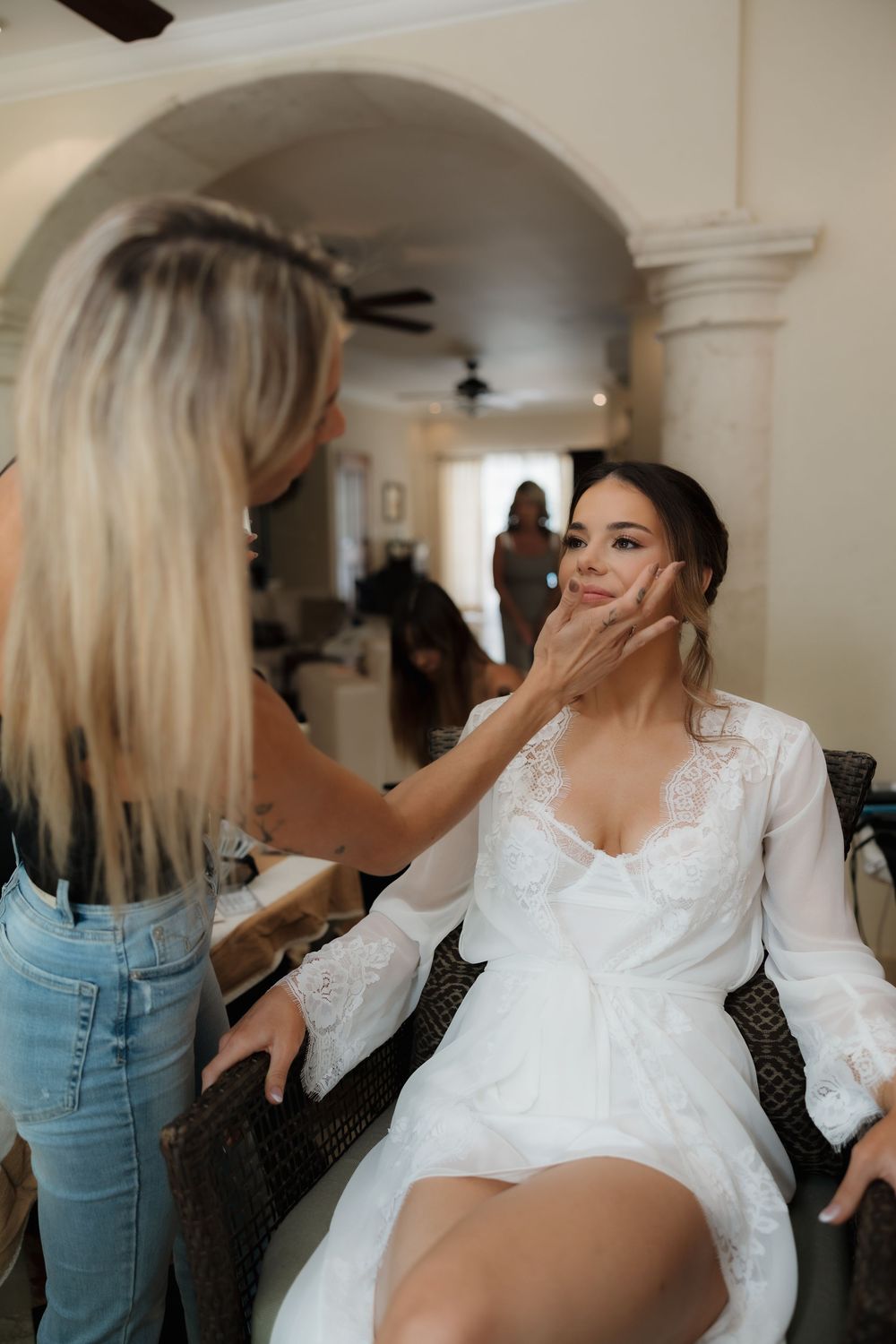 A bride in a white lace robe gets her makeup done by a stylist in jeans during wedding preparations.