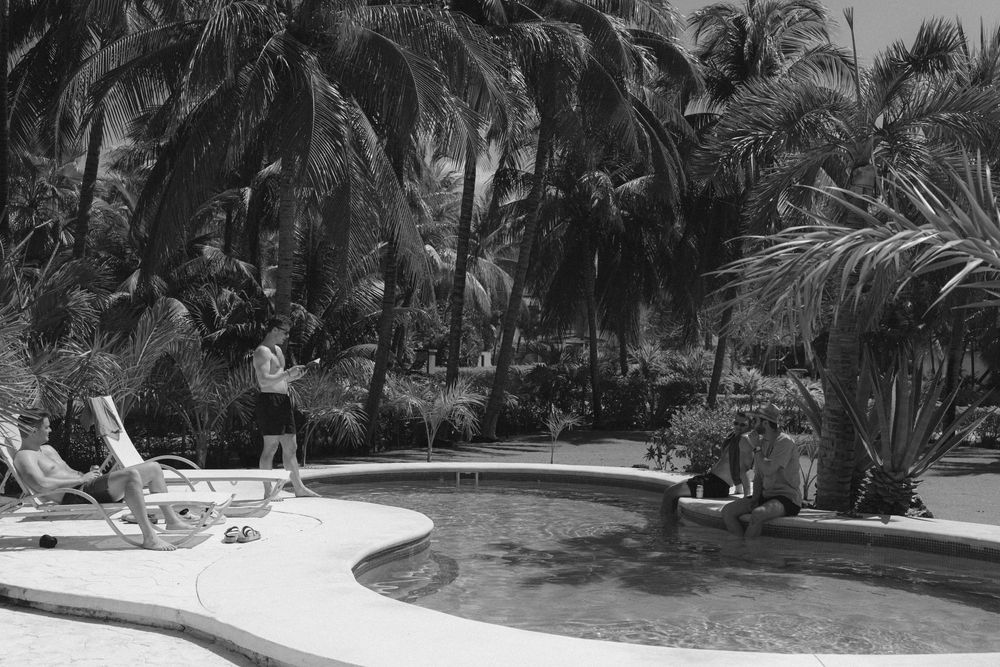 A vintage black and white photograph showing a curved swimming pool surrounded by tropical palm trees and lounge chairs.