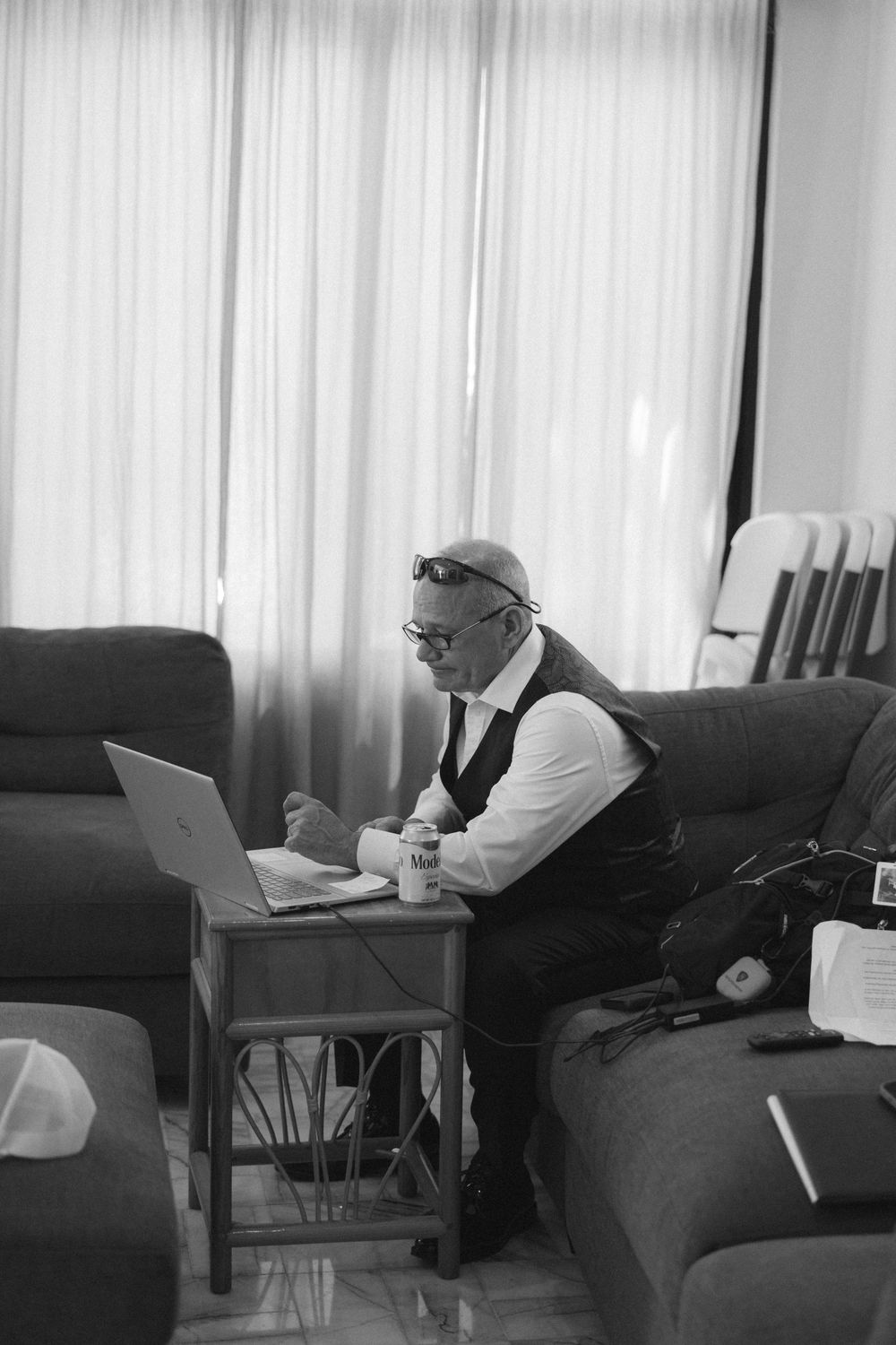 An elderly man in glasses works on a laptop while sitting on a couch in a sunlit room.
