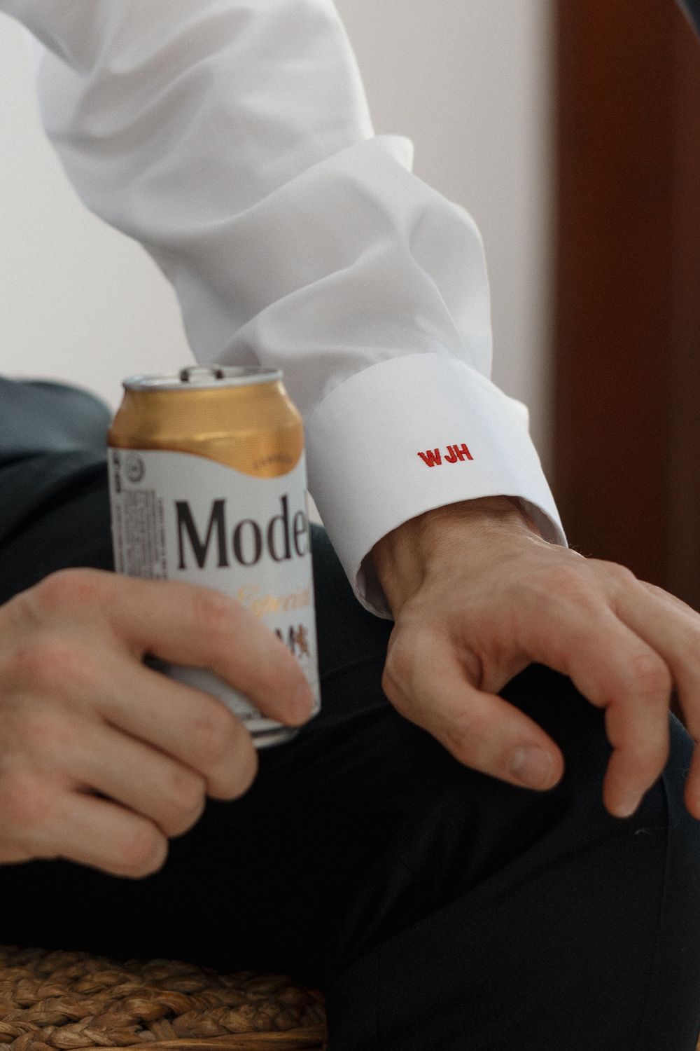 A hand wearing a white cuff with W&M initials holds a can of Modelo beer.