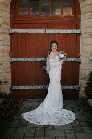 A bride in a lace wedding gown with long train poses in front of rustic wooden doors.