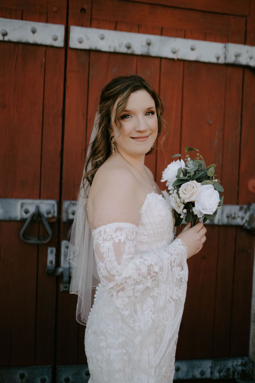Bride in a white lace wedding dress poses against a rustic red barn wall.