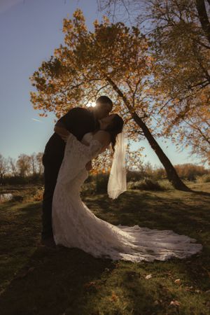 Silhouettes of a wedding couple embrace under autumn trees with sunlight streaming through branches.
