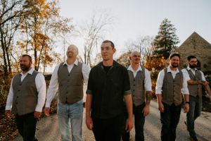 Groomsmen in gray vests and dark pants stand together at outdoor wedding venue.