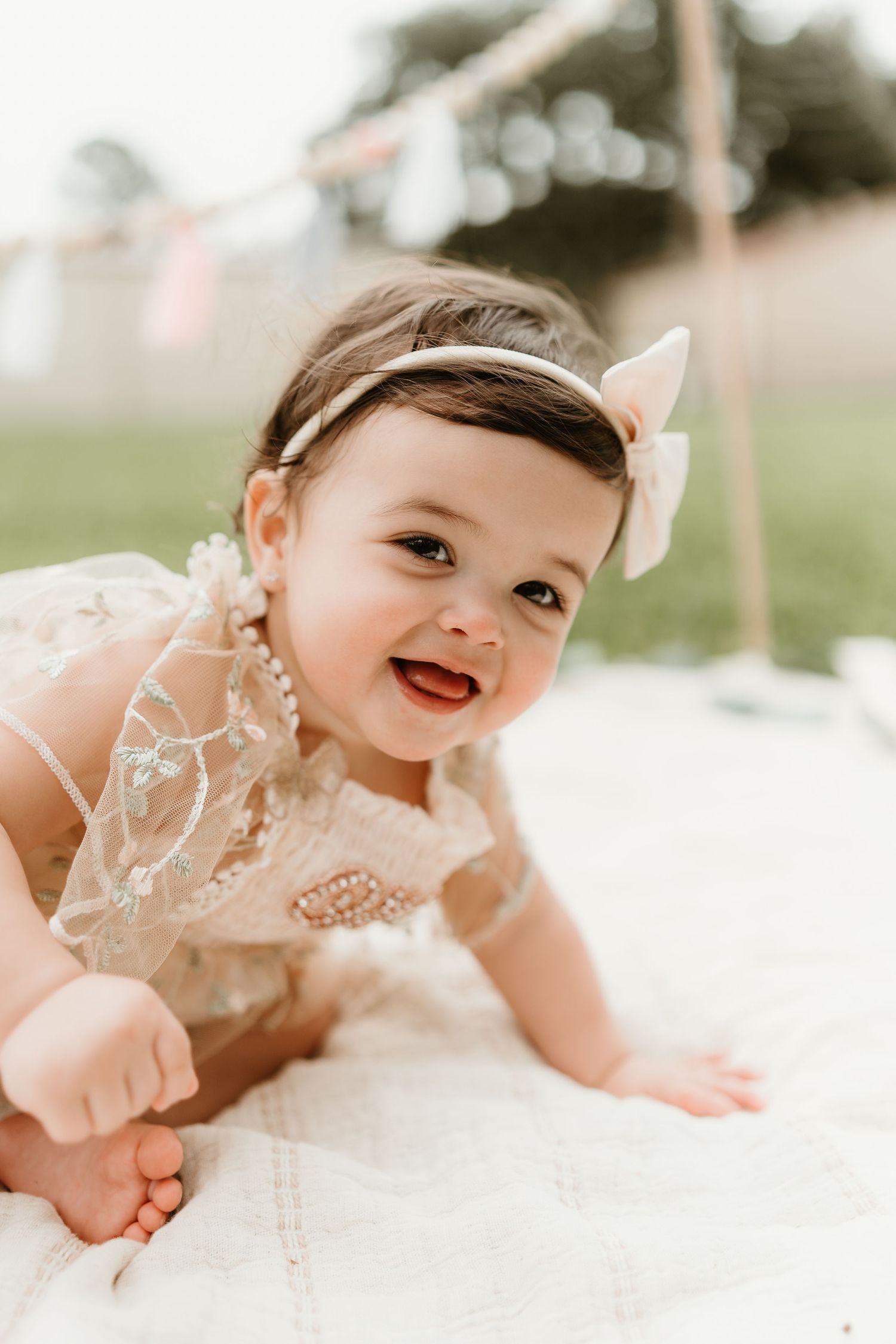 Baby in lace dress smiles while lying on white blanket during portrait session.