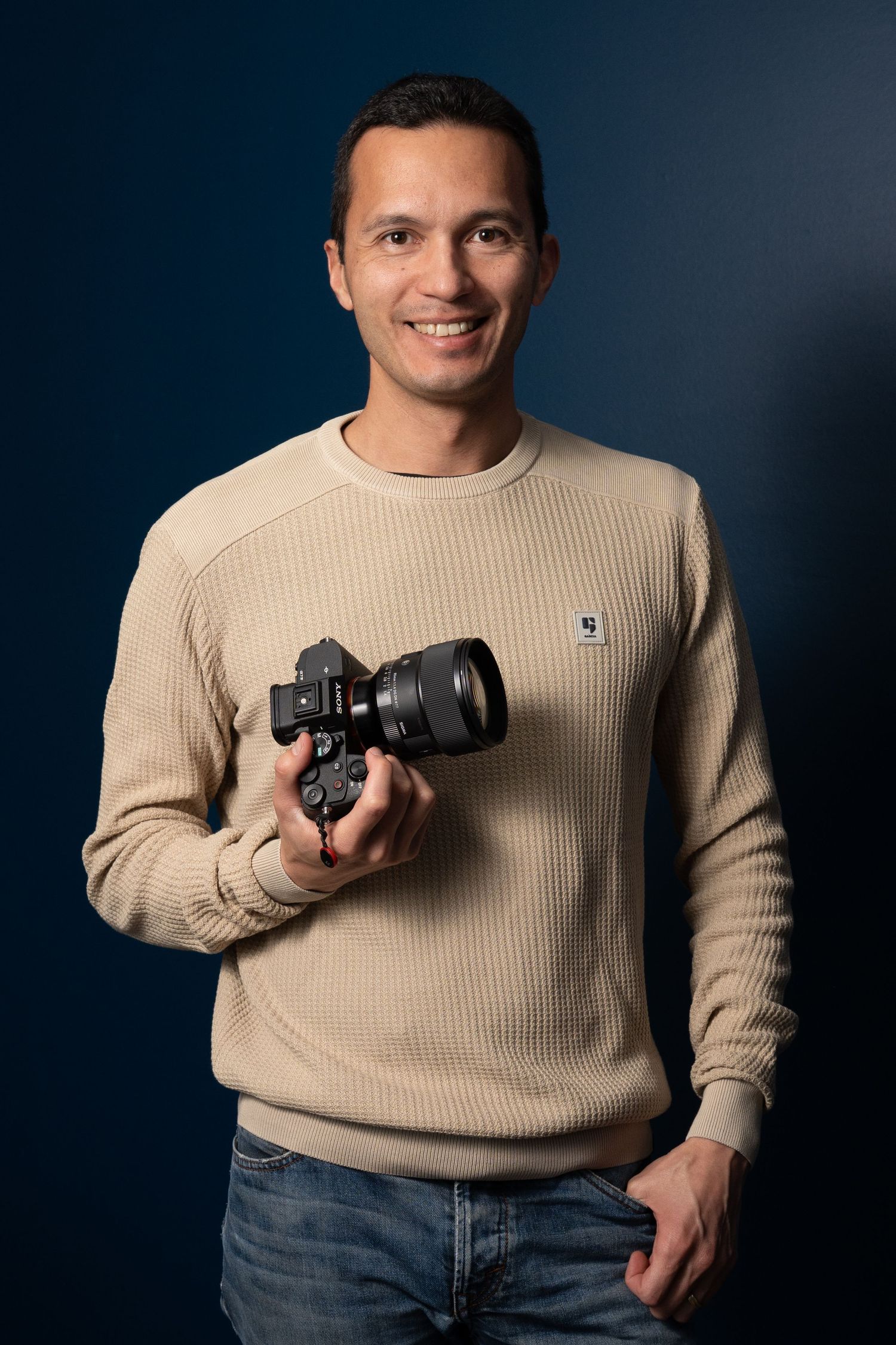 Photographe en pull beige tenant un appareil photo sur fond bleu foncé.