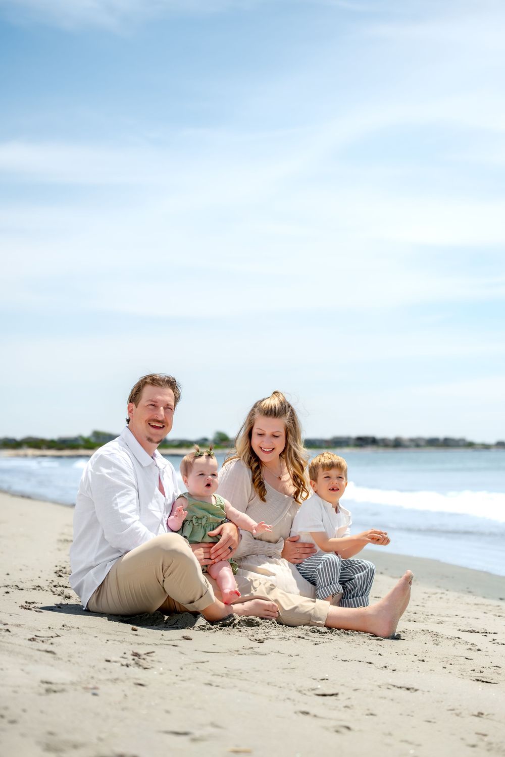 Family of four sitting on sandy beach, parents smiling with baby and toddler on sunny day.