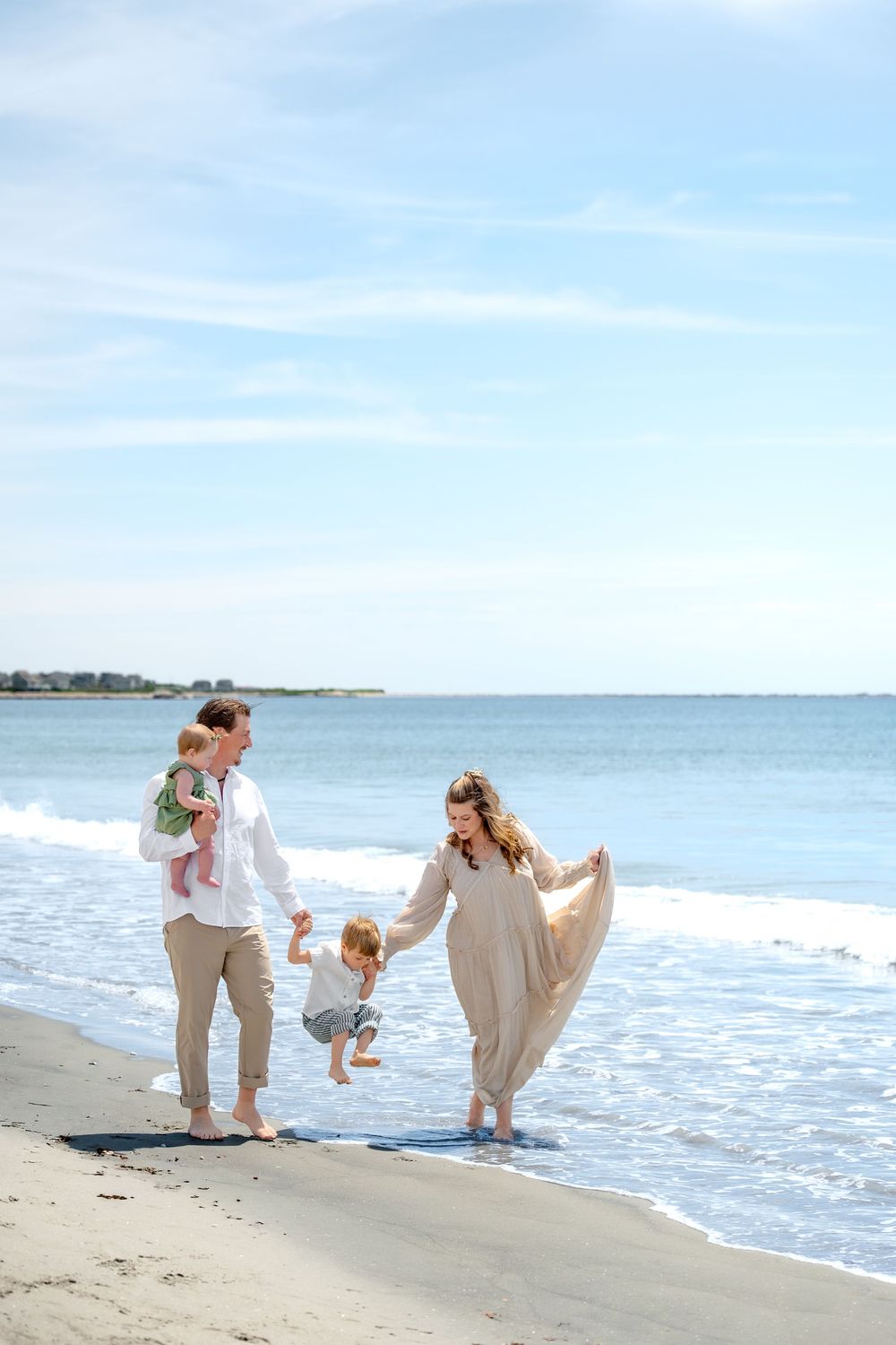 Family of four enjoying a sunny beach day, with parents swinging toddler over ocean waves while holding baby.