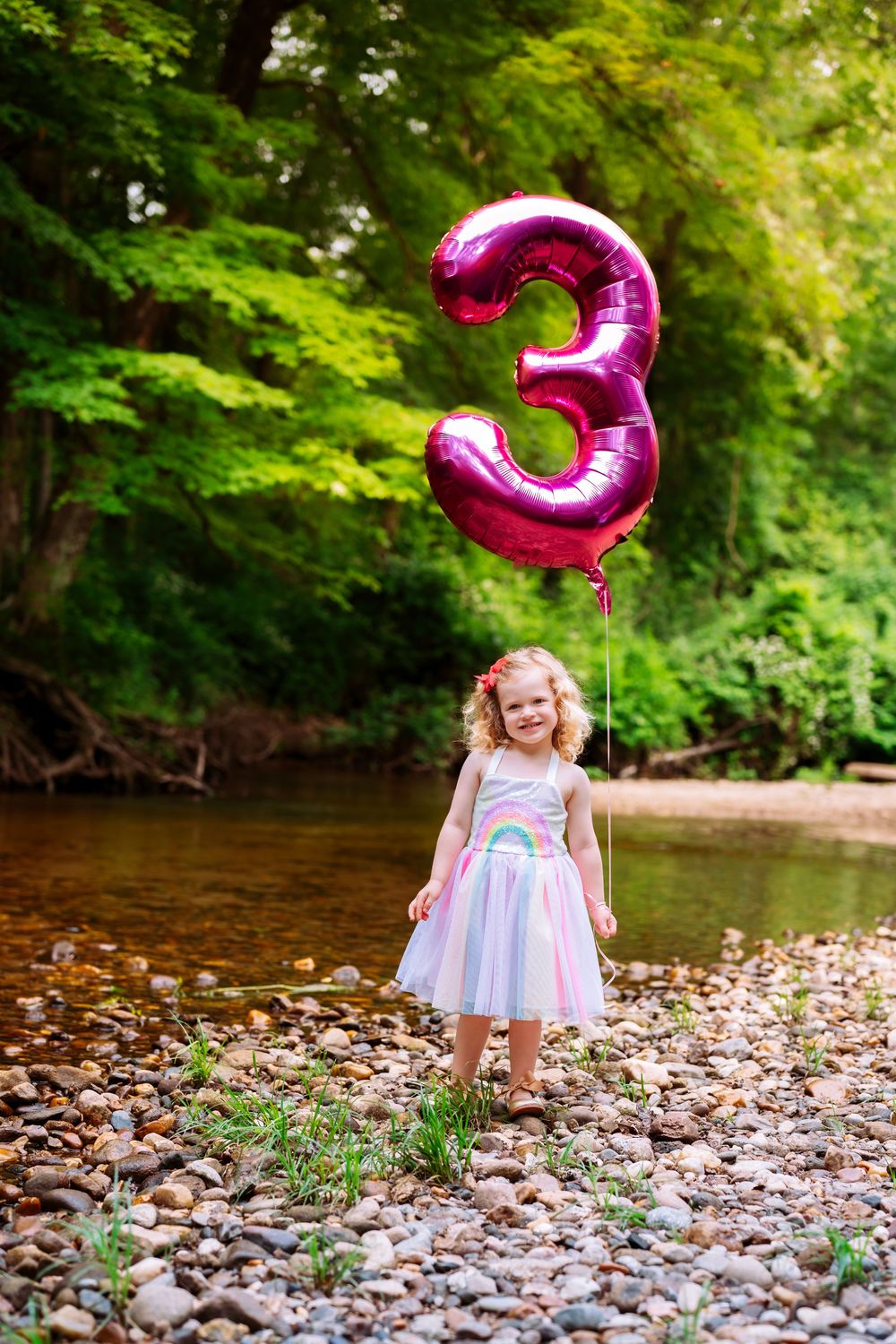 A pink number 3 balloon stands on a rocky riverbank surrounded by lush greenery.