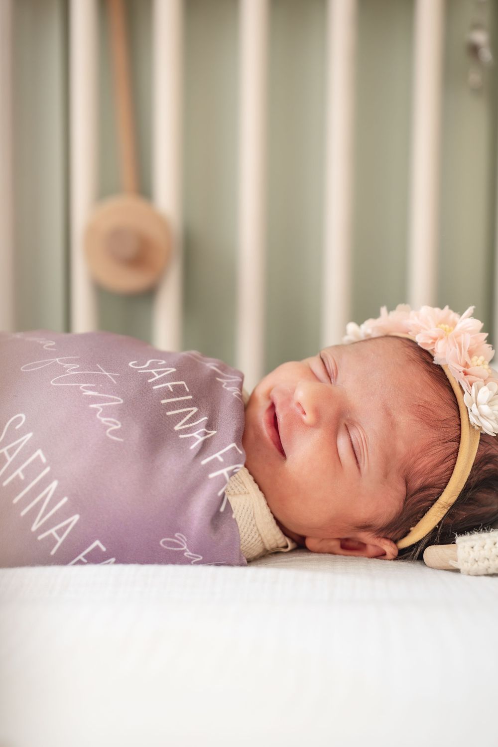 Sleeping newborn baby girl wrapped in personalized purple swaddle with floral headband in white crib.