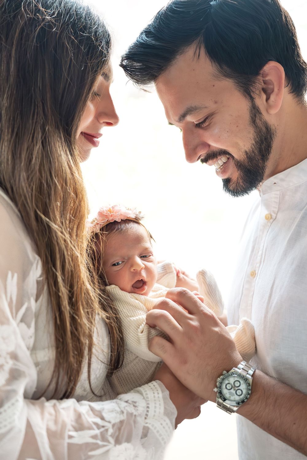 Happy parents smiling at their newborn baby wrapped in a cream blanket with a floral headband.