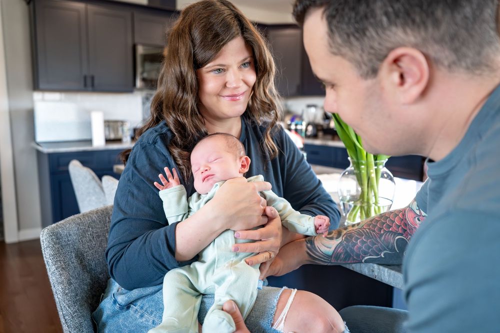 A woman holds a newborn baby while a tattooed man looks on lovingly in a modern home setting.
