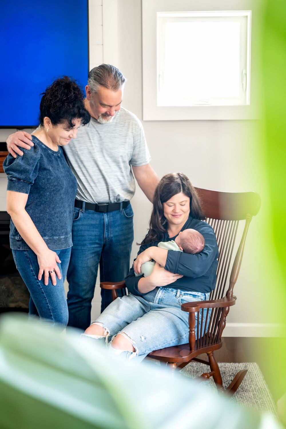 Family gathered around a newborn baby held by a woman in a rocking chair in a cozy home setting.