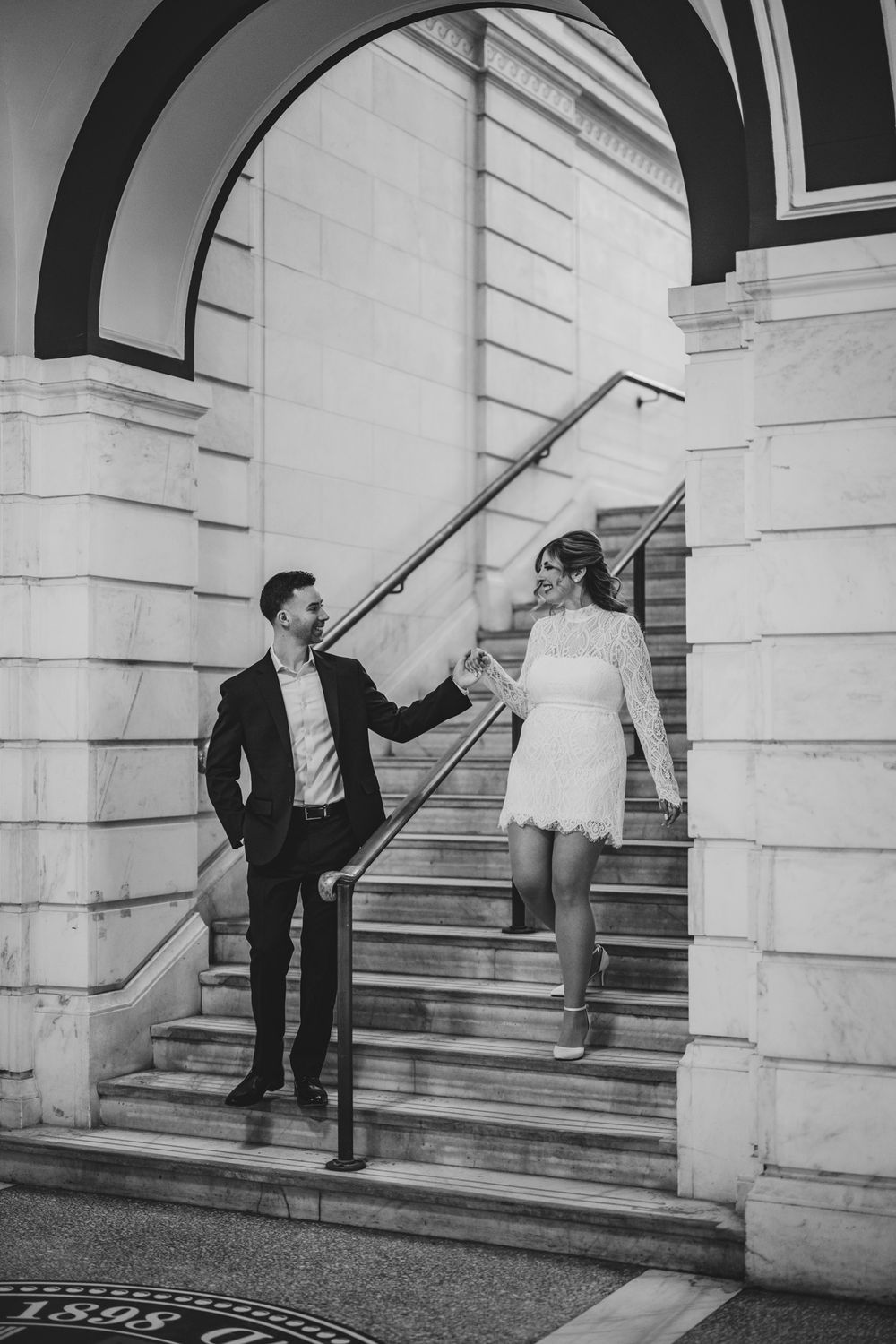 Black and white photo of couple posing playfully on grand marble staircase.