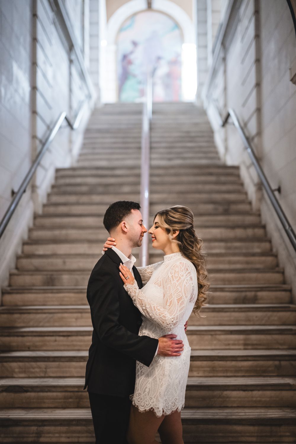 Couple in formal attire sharing romantic moment on white marble staircase.