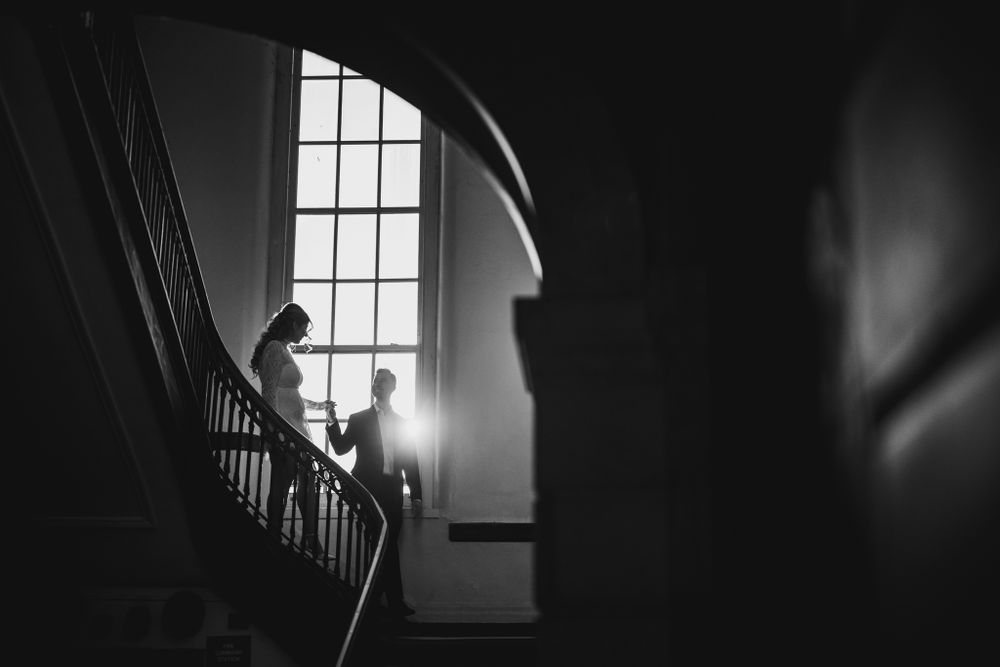 Moody black and white photo of figures in historic stairwell with window light.