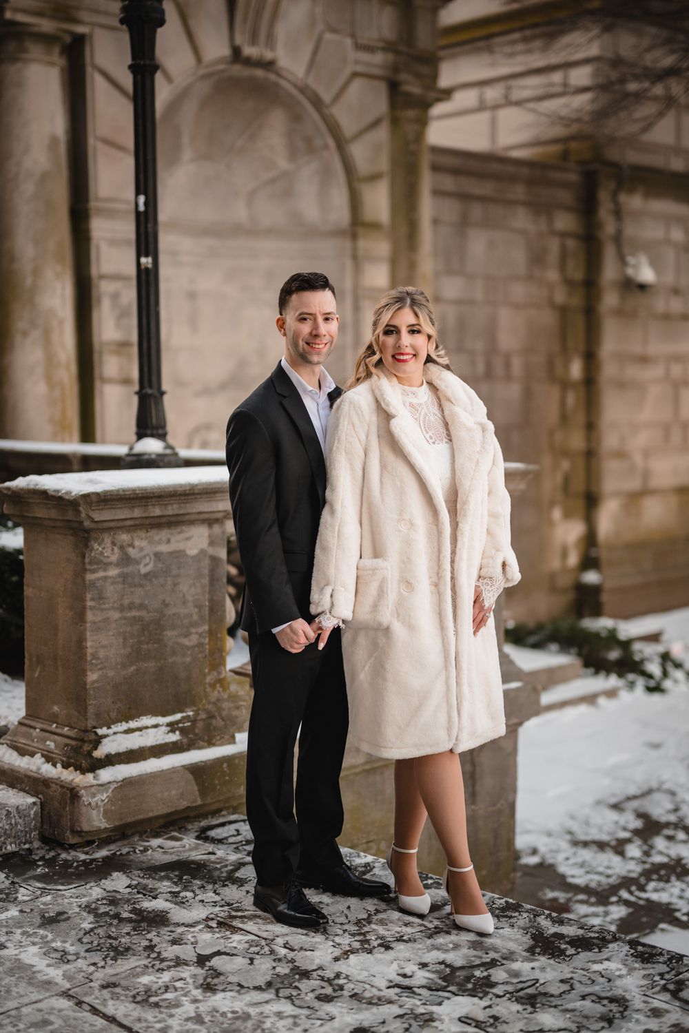 Winter wedding photo of couple in formal attire standing on snow-covered stone steps.