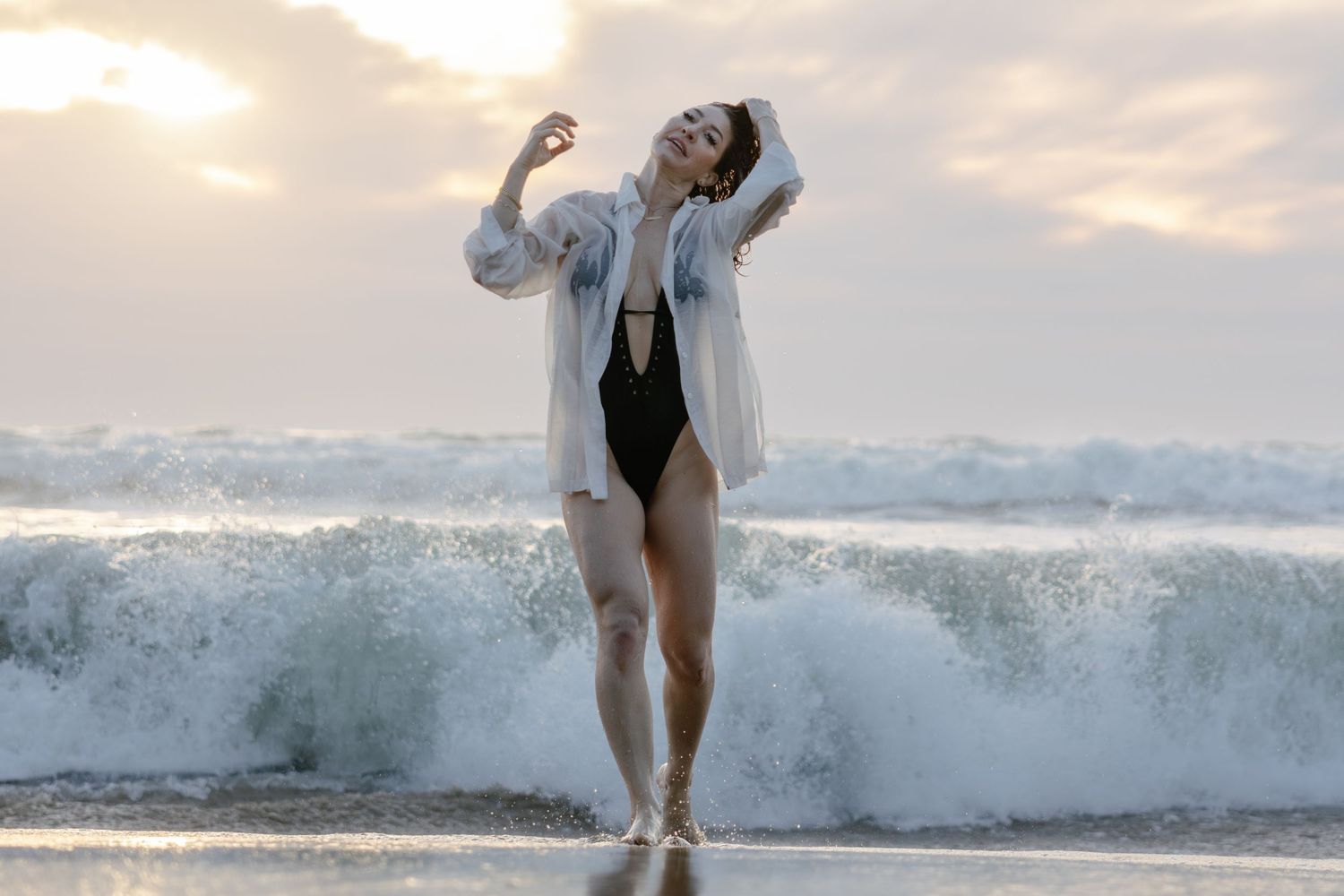 Someone in a white cardigan and black swimsuit dances joyfully on the beach as ocean waves crash behind them at sunset.