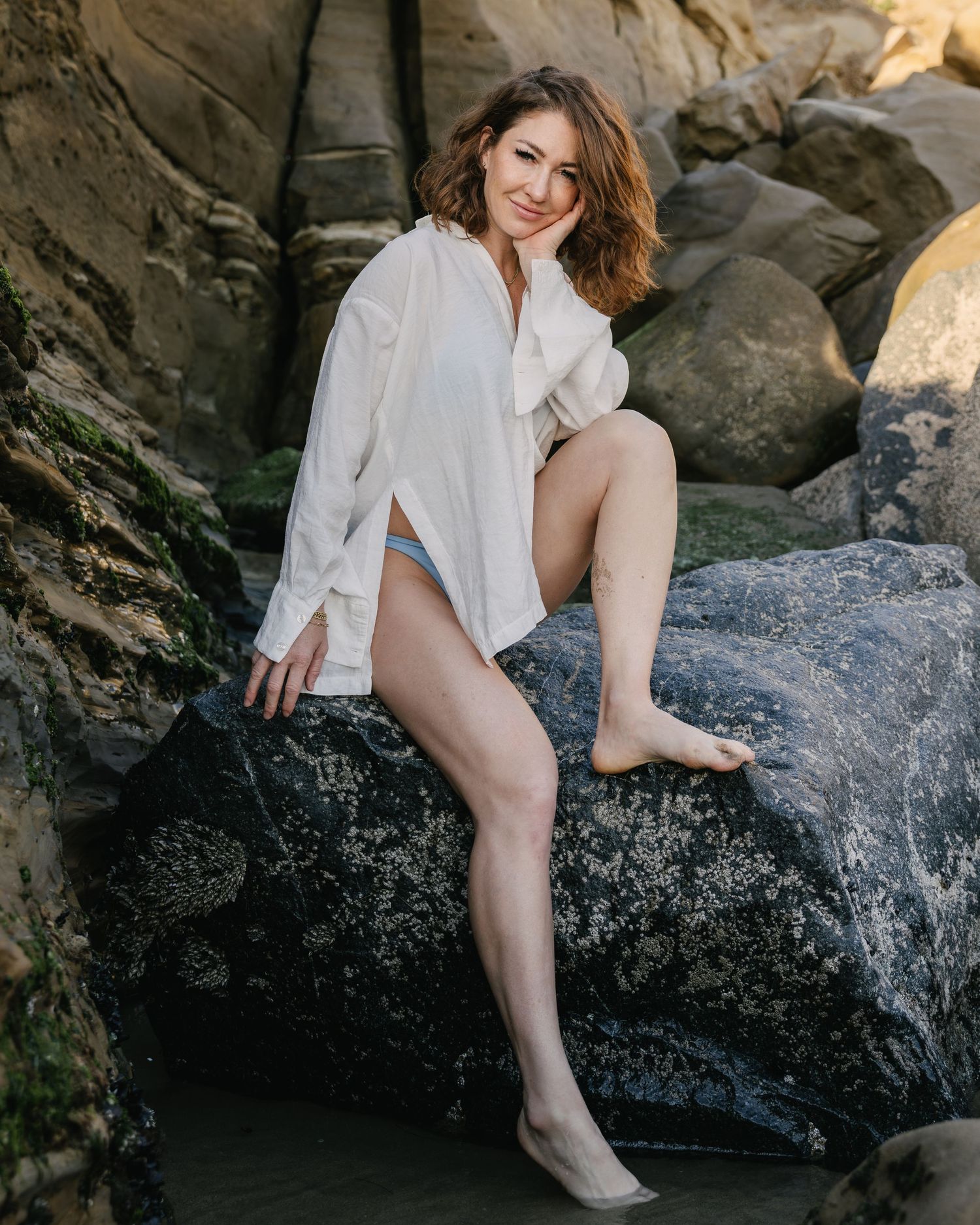 A white shirt model poses sitting on coastal rocks at sunset with warm natural lighting.